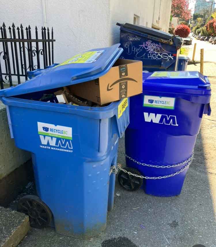 A recycling bin overflows with paper, including a large cardboard box which prevents the lid from closing.